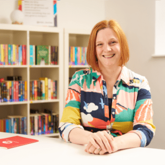 Jess - a white woman with shoulder length red hair - sits with her hands clasped in front of her, smiling at the camera. She wear a bright, colourful patterned shirt dress and is surrounded by colourful books.