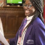 A young, teen, black girl with braided hair and purple school blazer stands smiling and proud after delivering a speech in UK parliament.