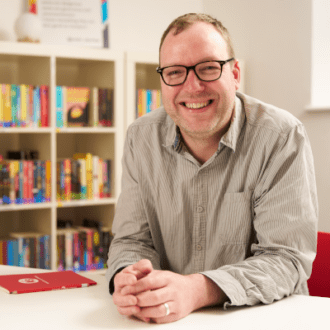Rob - a white man with short, fair hair - sits hands clasped in front of him at a white table. He has dark framed glasses and wears a relaxed, striped shirt. He smiles broadly at the camera surrounded by colourful books.