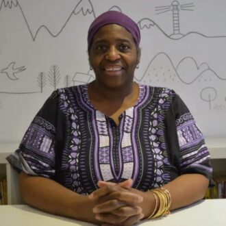 Sandra sits smiling with her hands intertwined on a white desk. She is wearing a deep purple and white patterned top with a purple head wrap.