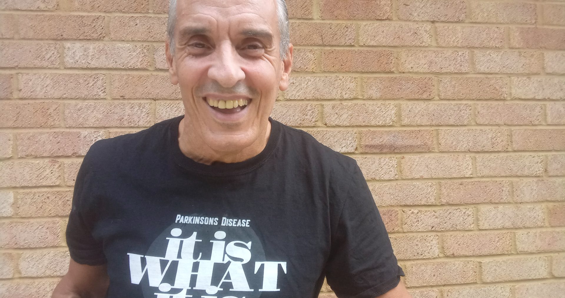 Joe smiles at the camera standing in front of a brick wall, wearing a Parkinson's Disease fundraising t-shirt