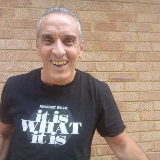 Joe smiles at the camera standing in front of a brick wall, wearing a Parkinson's Disease fundraising t-shirt
