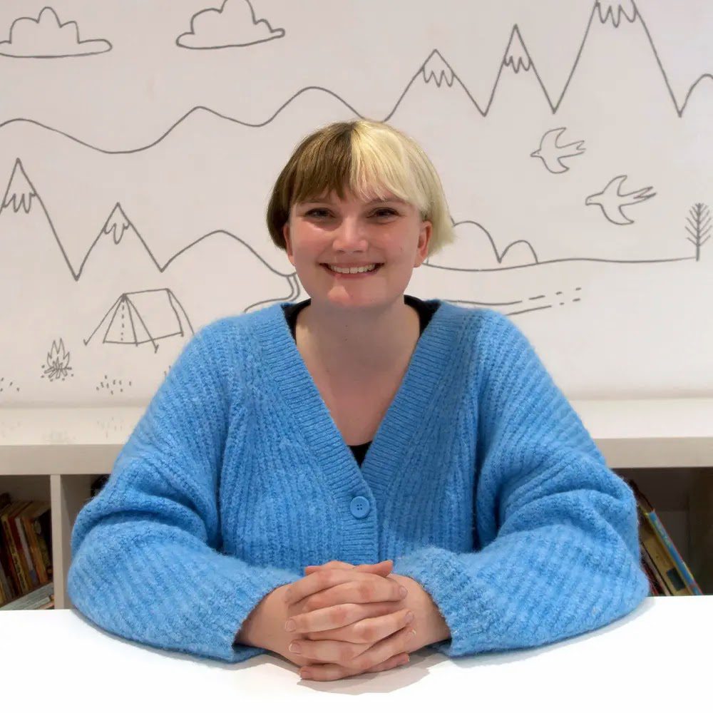 Rowan Earley sits smiling, hands clasped on a table in front of her. She has half blonde, half brown hair split down the middle. She wears a bright blue, oversized cardigan.