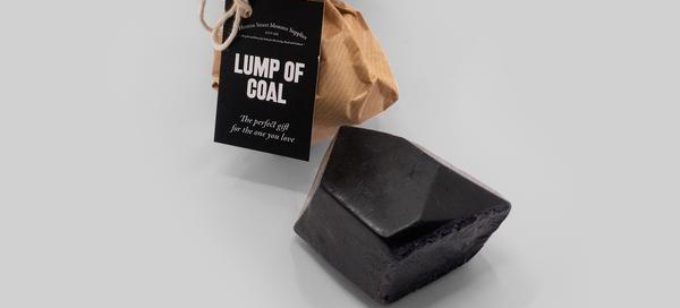 A black, shiny bar of soap in the shape of coal sits alongside brown wrapping paper, tied with string and black tag that reads 'Lump of Coal'