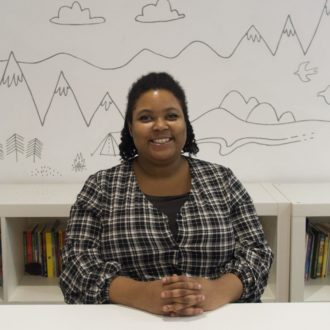 Maya Alexander has dark curly hair that stops just above her shoulders, she is wearing a black and white chequered shirt. Maya is smiling at the camera with her hands clasped in front of her on a white table.