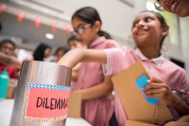 A smiling pupil reaches her hand into a silver bucket with a colourful sign on it that says 'Dilemmas'.