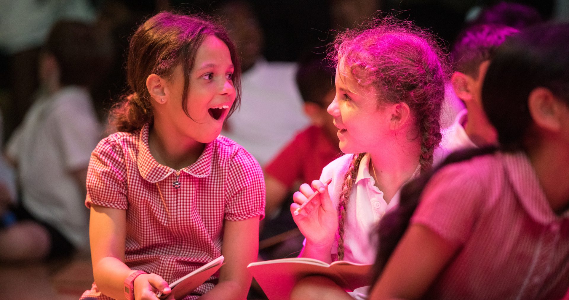 Two young girls, hair in plaits, wearing school uniform dresses, sit on the floor writing and excitedly exchange ideas