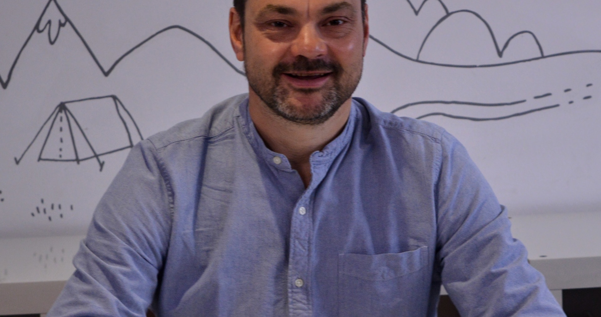 Justin wears a light blue shirt, has short dark hair and a short dark brown beard. He sits smiling with his hands clasped in front of him on a white desk.