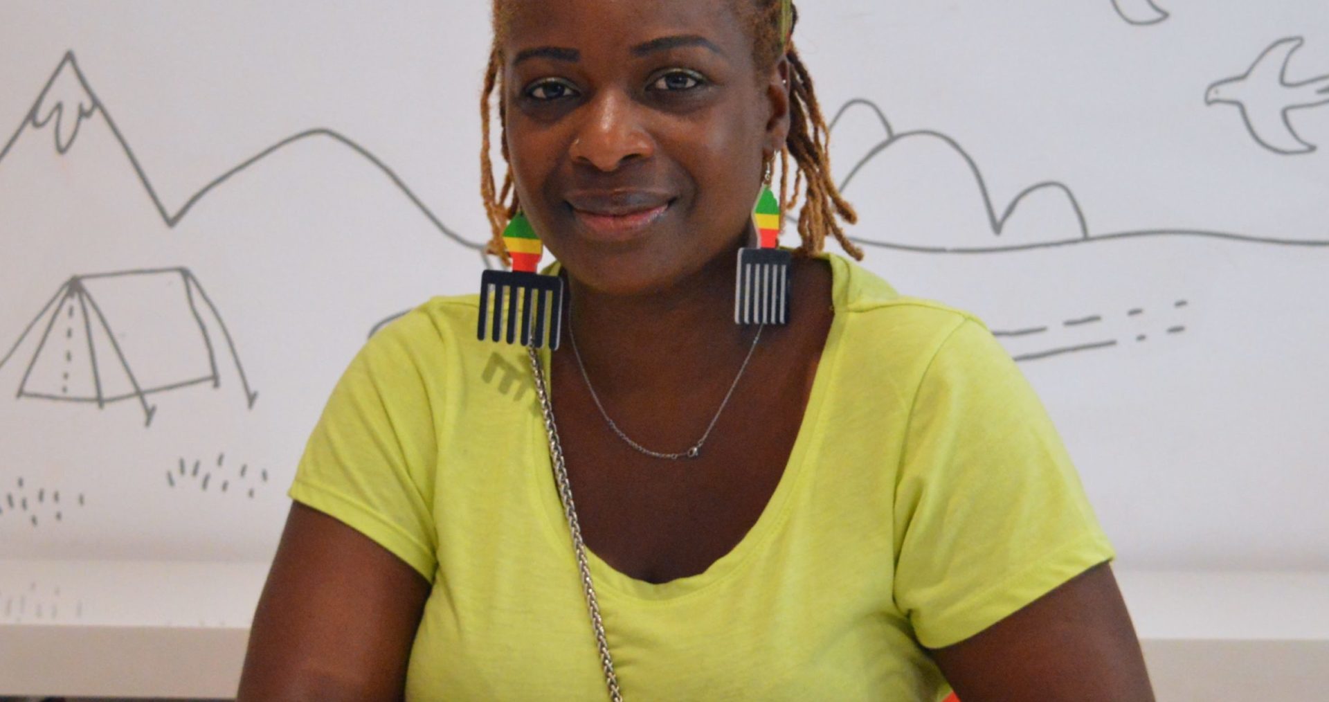 Shenelle has warm toned dreadlocks with a colourful scarf tied on top of her head. She has afro-comb earrings painted the colours of the Pan-African flag and is wearing a bright green top. She sits smiling at the camera with her hands clasped on the table in front of her. Her finger nails are colourful and sparkly.