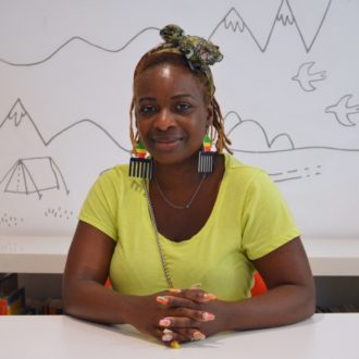 Shenelle has warm toned dreadlocks with a colourful scarf tied on top of her head. She has afro-comb earrings painted the colours of the Pan-African flag and is wearing a bright green top. She sits smiling at the camera with her hands clasped on the table in front of her. Her finger nails are colourful and sparkly.