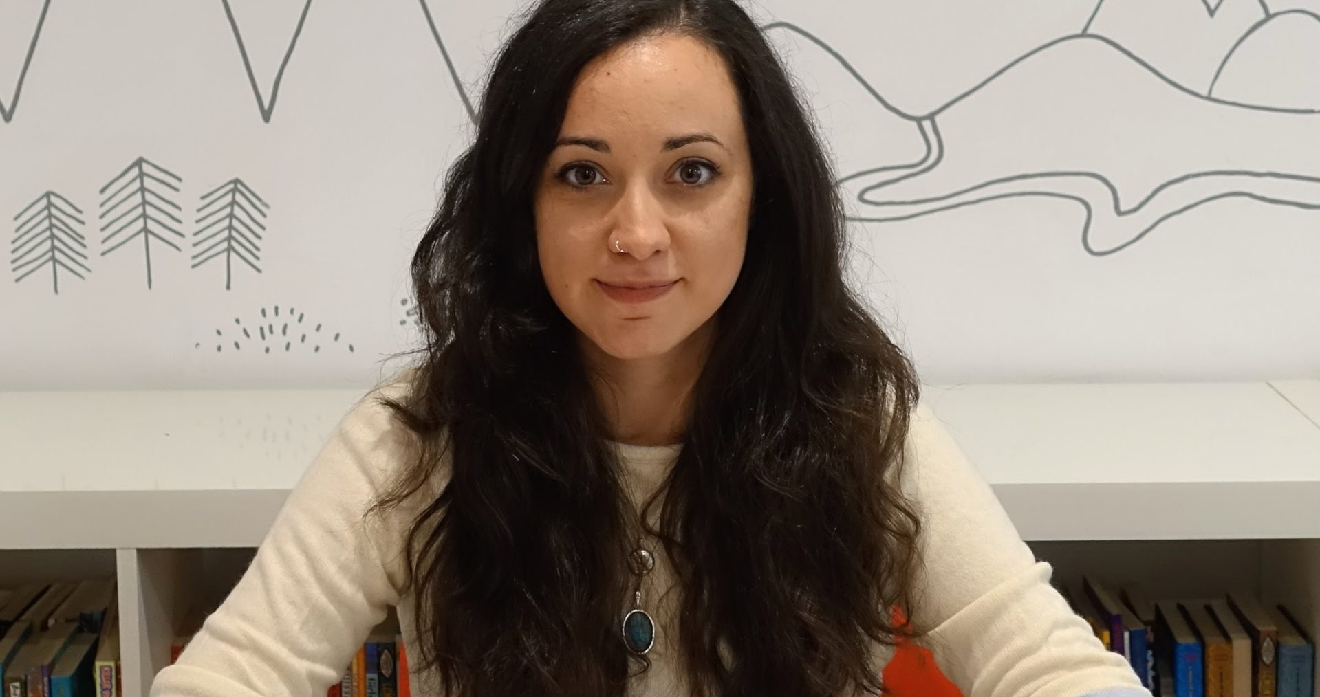 Alice has long dark, curly hair and is wearing a long sleeved white t-shirt. She has a silver nose ring, necklace and rings on. Her hands are clasped in front of her on the table and she is smiling at the camera.