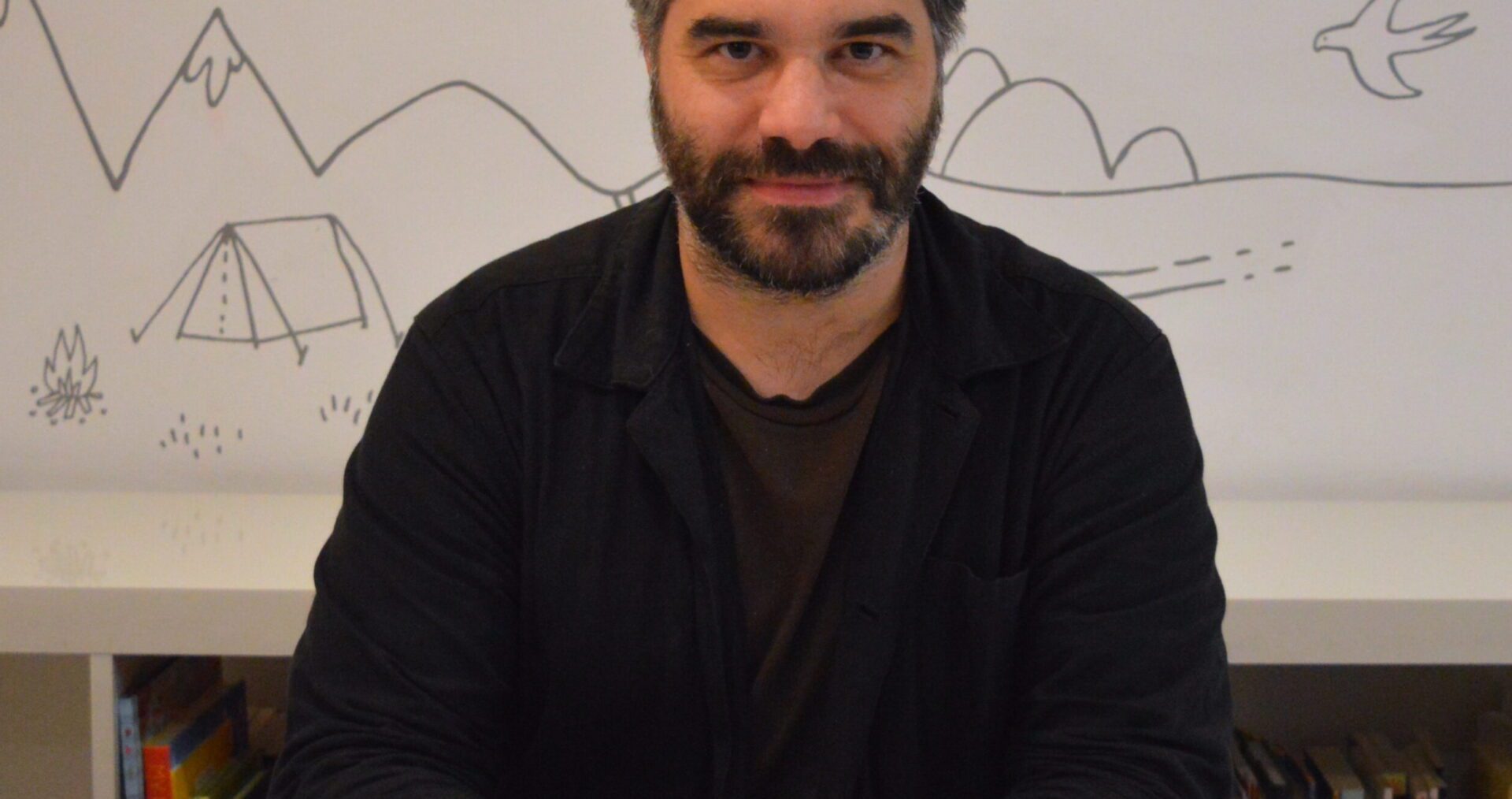 Michael Crowe sits with his hands folded on a white desk. He has short, styled black hair with flecks of grey above his ears. He has a short beard and is wearing a black jacket over a brown t-shirt.