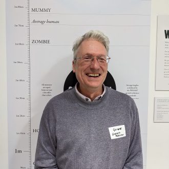 Gerard Darby stands smiling at the camera in front of a Hoxton Street Monster Supplies height chart. He wears a grey jumper and silver rimmed glasses.
