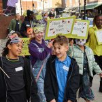 A group of young writers smile and laugh as they walk down Hoxton Street surrounded by placards and hand drawn monster masks