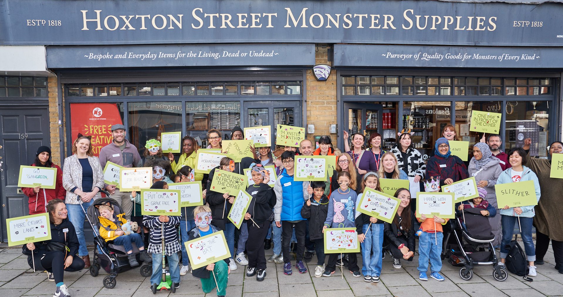 A large group of young people and adults stand in front of the grey Hoxton Street Monster Supplies shop waving lime green banners and placards with fun, monster puns written in black ink.