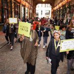 The group walks down the middle of Leadenhall Market, with its cobbled floor and ornate ceilings. The lift their lime green placards above their heads.
