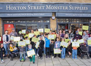 A large group of young people and adults stand in front of the grey Hoxton Street Monster Supplies shop waving lime green banners and placards with fun, monster puns written in black ink.