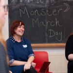Dorothy, wearing a dark blue top and smiling, sits in front of the Ministry blackboard which has Monster Month chalked on it