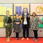 Four members of the Ministry of Story staff, dressed to impress, stand smiling and pulling expressive, happy poses in front of a large, colourful Ha Ha Hackney poster
