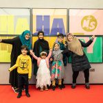 A large group of mums and children, dressed to impress, stand smiling and pulling energetic and fun poses in front of a large, colourful Ha Ha Hackney poster