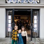 Three young children stand with there arms around each other in front of the entrance to Curzon cinema in Hackney