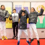 Four young teenage boys, wearing white jeans and puffer jackets jump up high in front of a large, colourful Ha Ha Hackney poster