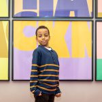 A young black boy in a blue and orange stripy jumper poses proudly in front of a super sized, colourful Ha Ha Hackney poster