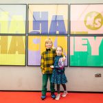 Two young children, dressed in sequins and a yellow jacket, stand smiling stand and proud with their arms around each other in front of a large, colourful Ha Ha Hackney poster
