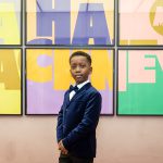 A young black boy dressed in a blue velvet suit jacket and bow tie, stands proud and happy in front of a large, colourful Ha Ha Hackney poster