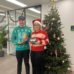 Two smiling people stand in front of a Christmas tree holding a tin of Bah! Humbugs