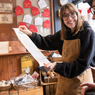 Lina is a white woman, with shoulder length brown hair and a fringe. She wearing large glasses, a shopkeepers apron and has been photographed holding a shop product. She is smiling broadly.