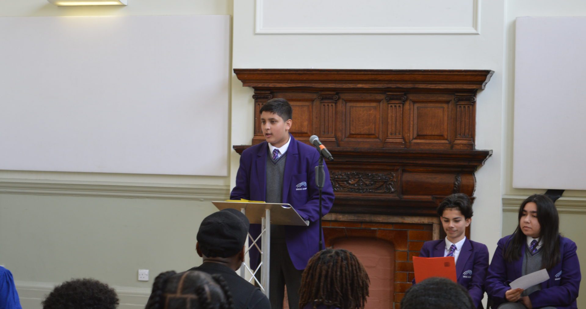 A young teenager stands in a purple school blazer delivering a speech to the audience at Shoreditch Town Hall.