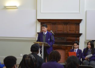 A young teenager stands in a purple school blazer delivering a speech to the audience at Shoreditch Town Hall.