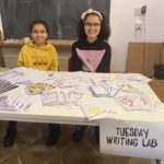 Two young writers smile sitting behind a table piled high with their hand created zines.