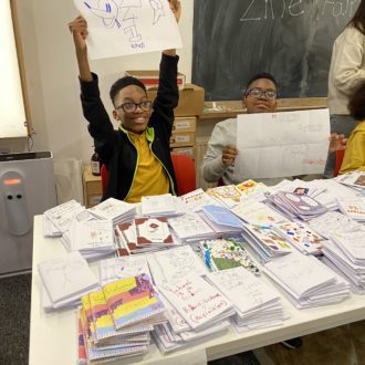 A joyful young writer in a yellow t-shirt holds their zine up above their head showing it proudly to the photographer