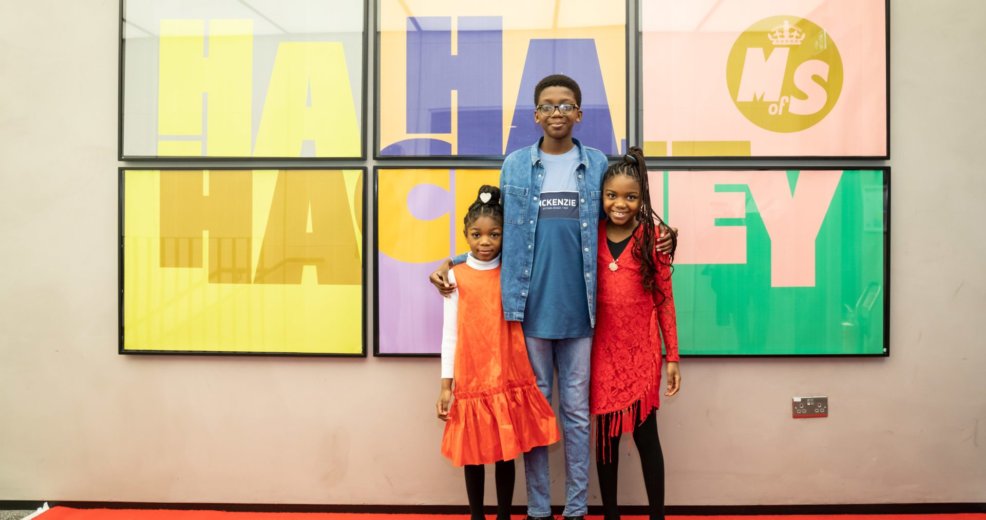 Three young people, dressed in bright clothing, stand smiling, arms around each other, in front of a large cinema poster that reads Ha Ha Hackney with the Ministry of Stories logo