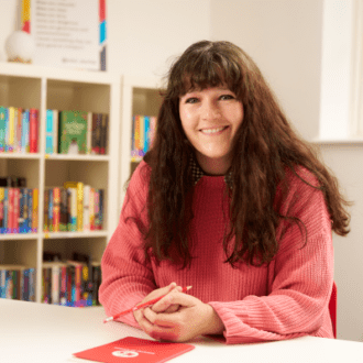 Jenny - a white woman with a fringe and long, dark and wavy hair - sits with her hand clasped in front of her. She wears a coral pink jumper and smiles happily at the camera.