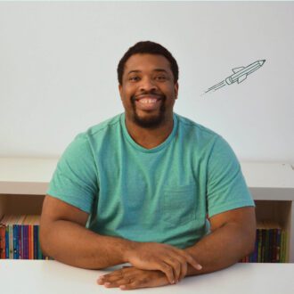Mayo sits smiling broadly with his hands folded on top of a white desk. He is a black man, with short curly black hair and a goatee. He is wearing a green t-shirt.