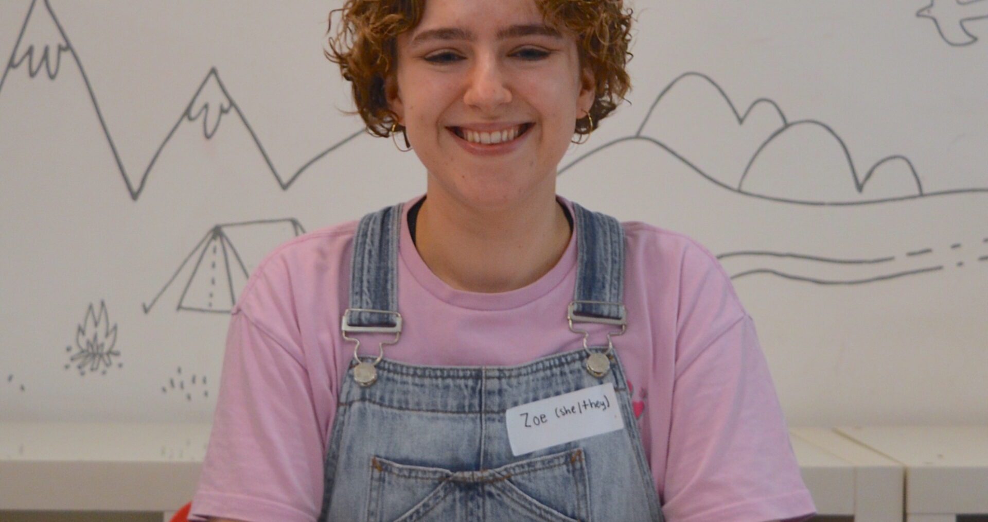 Zoe sits with their hands clasped smiling at the camera. She is a white woman with short curly hair, gold hoop earrings and a watch on her right wrist. They are wearing a lilac purple t-shirt and light blue denim dungarees.