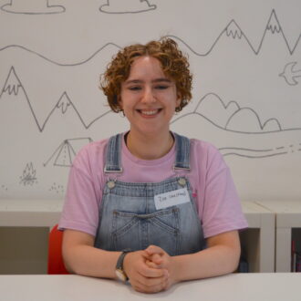 Zoe sits with their hands clasped smiling at the camera. She is a white woman with short curly hair, gold hoop earrings and a watch on her right wrist. They are wearing a lilac purple t-shirt and light blue denim dungarees.