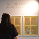 A young woman - with long brown hair, black top and grey skirt -looks at the writing set out on the wall. We see her from the back and she is silhouetted by the exhibition spotlights. The writing is on small pieces of white paper, framed against gold paper. There are two frames, each with four piece of writing displayed.