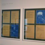 A bright, white wall displays writing on small pieces of white paper, framed against gold paper accompanied by two cyanotypes illustrations of blue and white that depict young faces
