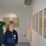 A young boy with blonde hair and blue jumper smiles shyly at the camera as he stands by the white wall displaying his writing