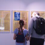 A young girl - with her hair tied in a high ponytail and wearing a small black backpack - stands with her dad - bald, wearing a black backpack and grey long sleeve top - look at the writing on the wall. We see them from the back as they are silhouetted by the exhibition spotlights. The writing is on small pieces of white paper, framed against gold paper. There are three frames, each with four piece of writing displayed.