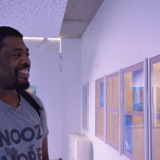 A young black man wearing a white t-shirt smiles broadly while looking at the writing displayed on the wall in front of him