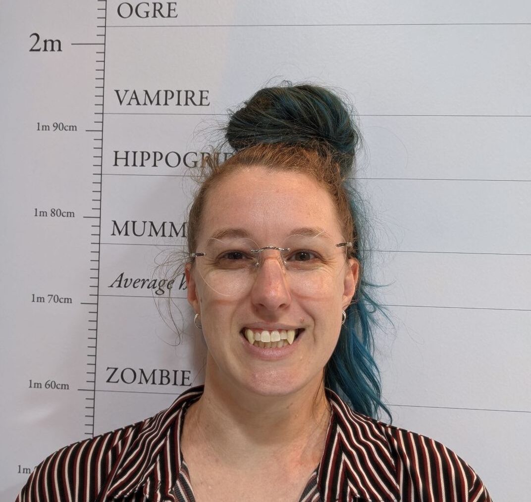 Claire stands smiling in from of a height chart depicting different monsters, the height marker indicates that they are a vampire. They have long blue hair tied up in a bun on top of their head, they're smiling at the camera with fake fangs poking out of their mouth. Claire is a white person, wearing a striped brown and white shirt and rimless glasses with lenses shaped like love hearts.