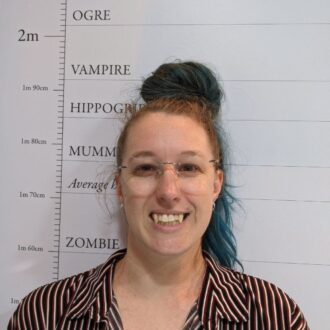 Claire stands smiling in from of a height chart depicting different monsters, the height marker indicates that they are a vampire. They have long blue hair tied up in a bun on top of their head, they're smiling at the camera with fake fangs poking out of their mouth. Claire is a white person, wearing a striped brown and white shirt and rimless glasses with lenses shaped like love hearts.