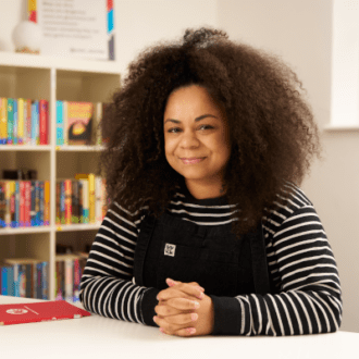 Sabrina - a black woman with dark, curly hair - sits with her hands clasped in front of her. She wears a long sleeved striped top and navy dungarees. She smiles happily a the camera.