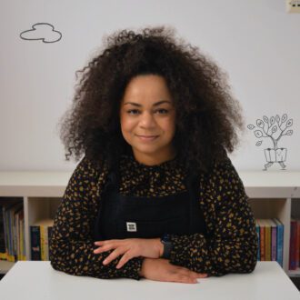 Sabrina sits smiling warmly with her arms folded on a white desk. She is a black woman with afro curly brown hair. She is wearing a black dungaree dress over a black shirt patterned with small orange flowers.