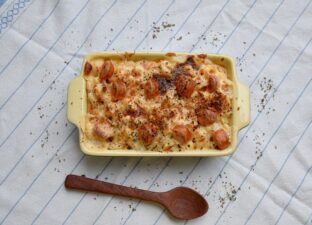A full and tasty lasagna, bubbling with cheese and tomato, sits in a traditional, cream oven dish in the centre of a table. The table is covered in a white linen cloth with blue strips. Next to the oven dish is a brown, wooden cooking spoon and a few sprinkles of black pepper.